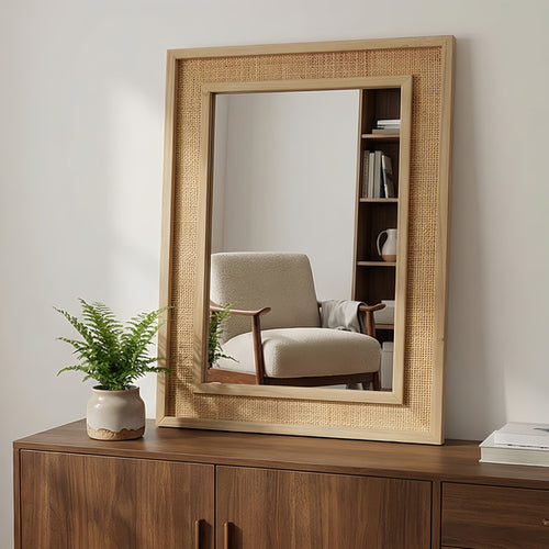 Rectangular mirror with wicker frame on a wooden dresser, reflecting an armchair and bookshelf.