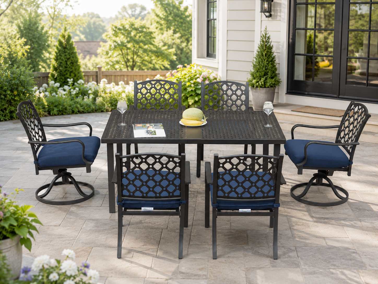 A 7-piece outdoor patio dining set featuring a rectangular black metal table and four matching swivel chairs with blue cushions, set on a stone patio in front of a house.